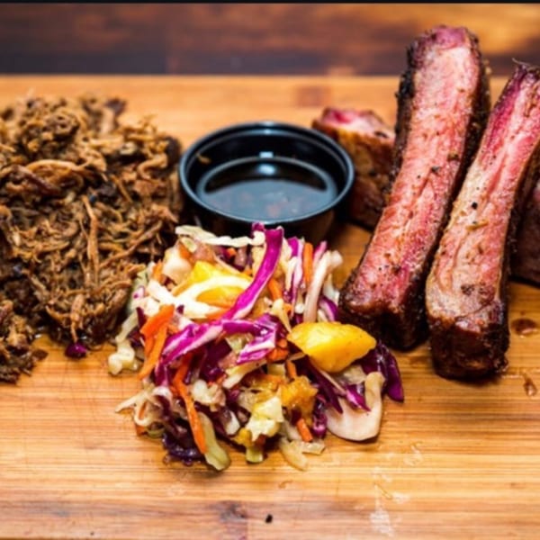 The Taste Smokers' BBQ platter with pulled pork, ribs, coleslaw, and a side of sauce on a wooden board.