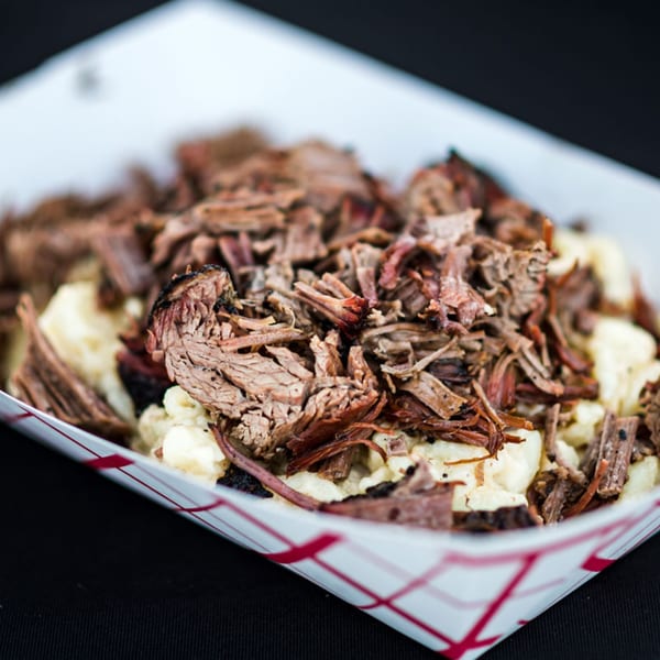 Close-up of The Taste Smokers' pulled brisket over creamy mac and cheese in a paper tray.