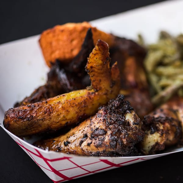 Close-up of The Taste Smokers' BBQ chicken wings, fries, and cornbread in a paper tray.